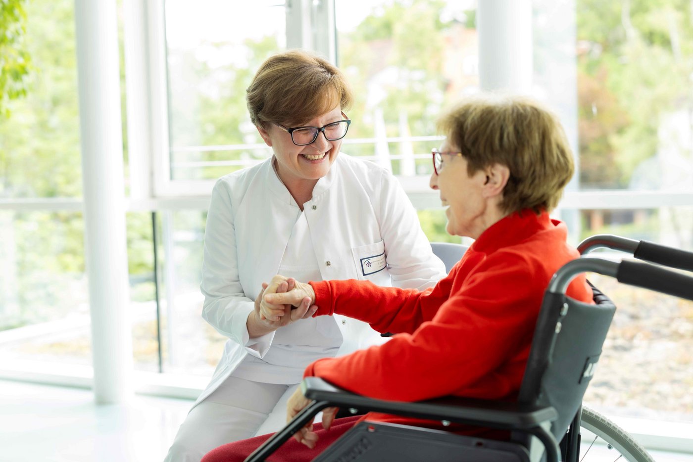 Eine Ärztin im Arztkittel interagiert mit einer älteren Patientin im roten Bademantel, die im Rollstuhl sitzt